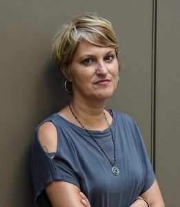 Photo of Kelcey Parker Ervick. She is standing against a wall, wearing a blue shirt.